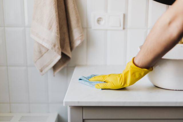 Yellow-gloved hand wiping a white bathroom counter with a blue cloth.